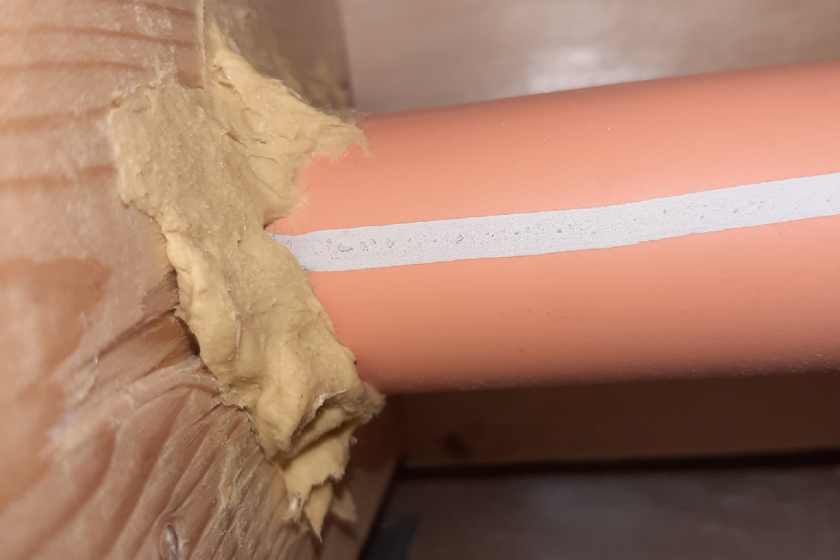 An orange CPVC fire sprinkler pipe passing through a wall penetration, with a bead of white caulk applied where the pipe meets the yellow fiberglass insulation