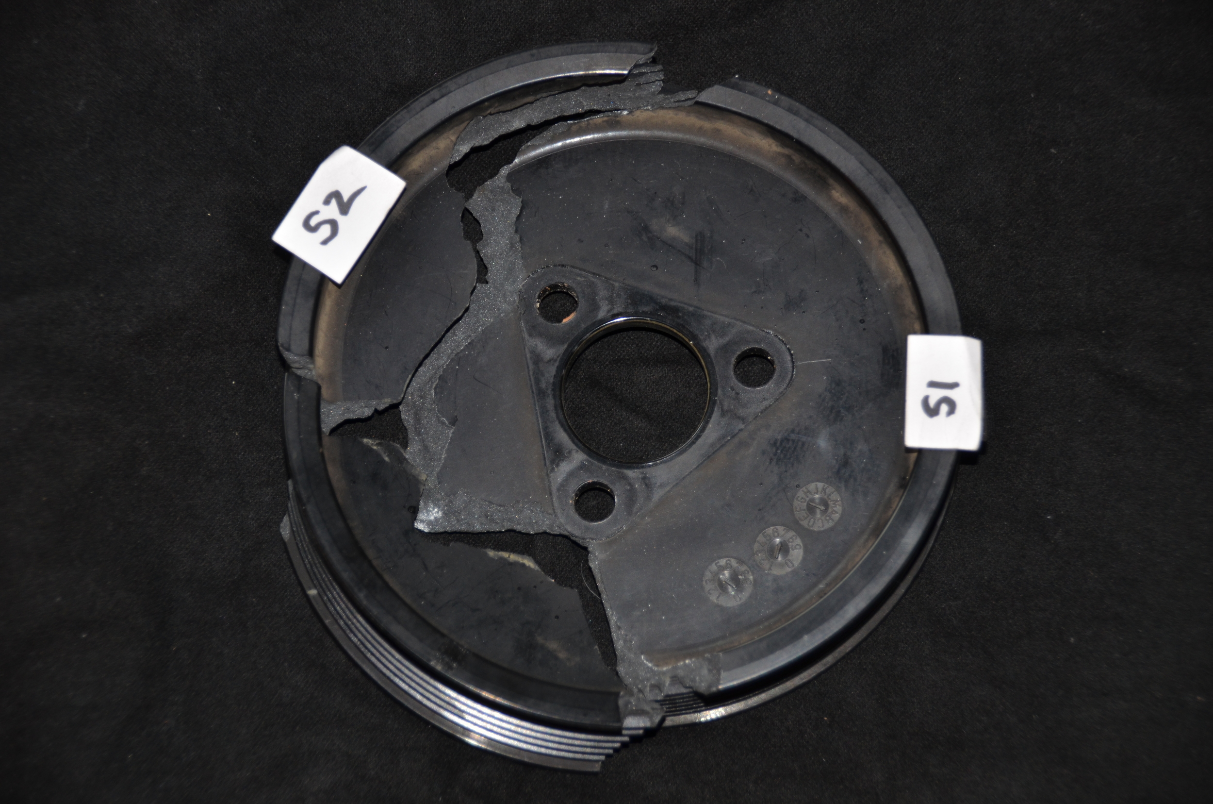 The fractured black composite automotive pulley photographed on dark evidence cloth with paper exhibit labels S1 and S2, showing a large wedge-shaped section broken out of the pulley face