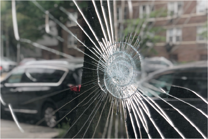 A transparent panel shattered by an impact, with long radial cracks spreading from a central impact star, photographed against a street of parked cars