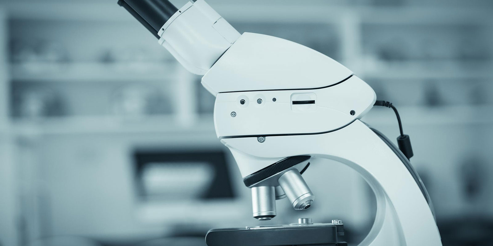 A white laboratory optical microscope on a bench, photographed against a blurred laboratory background