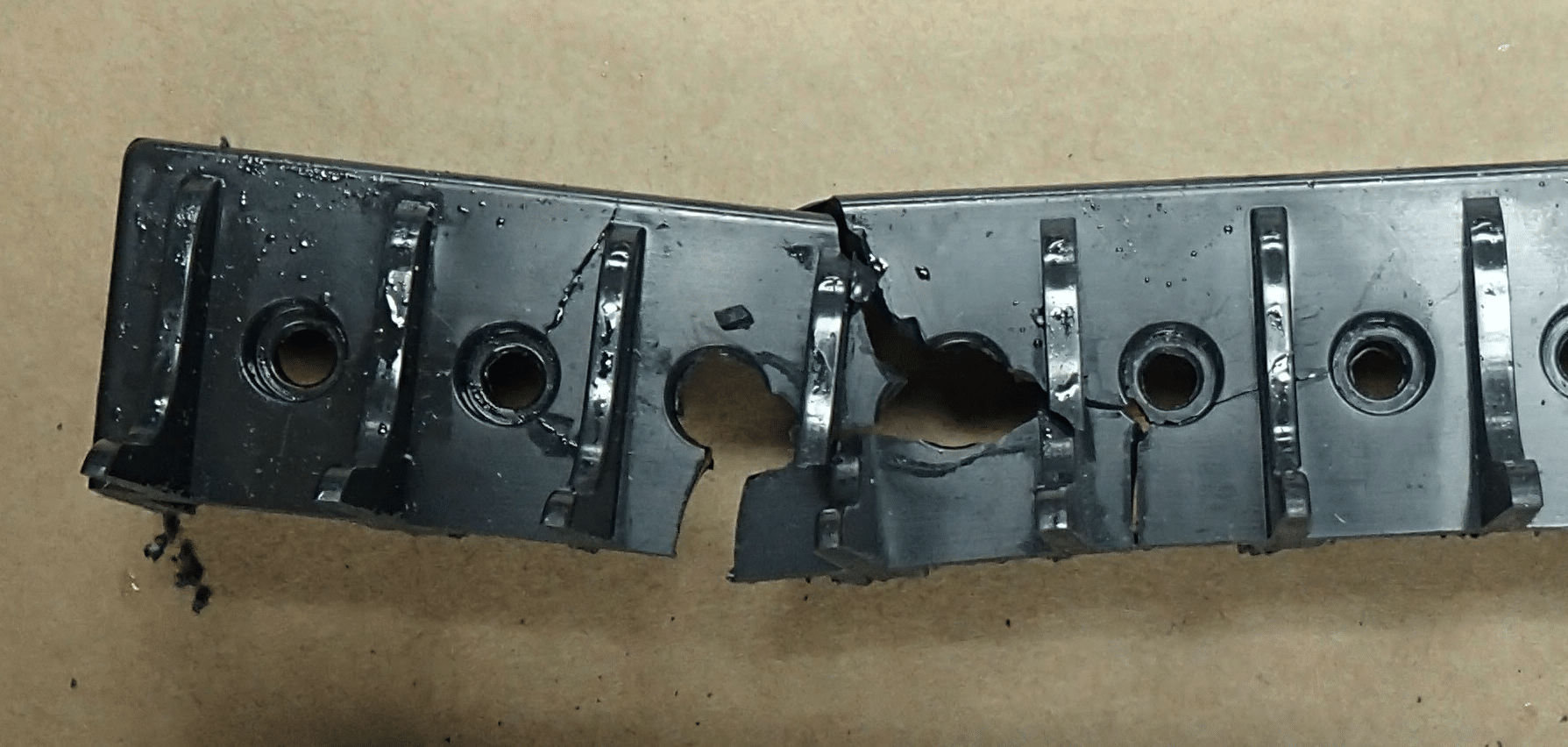 A sectioned black plastic assembly with a row of metal-lined ports, showing a large irregular fractured opening and loose debris where material has broken away