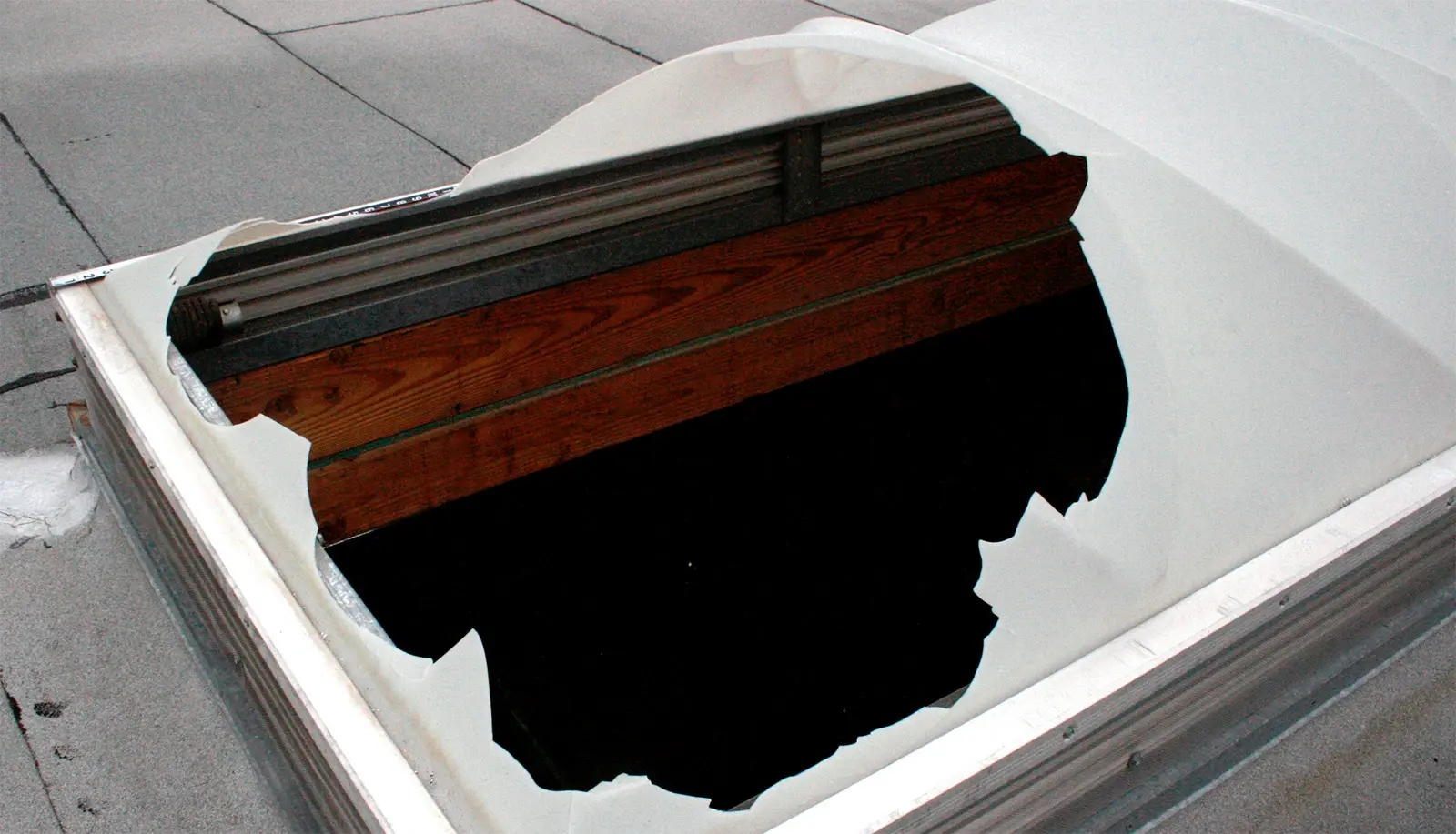 A white domed plastic skylight on a rooftop with a large jagged hole broken through it, exposing the dark roof interior and timber below
