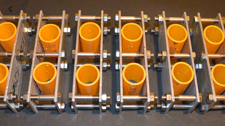 Two rows of orange CPVC pipe rings held under stress in bolted stainless steel fixtures on a dark bench, with handwritten specimen markings