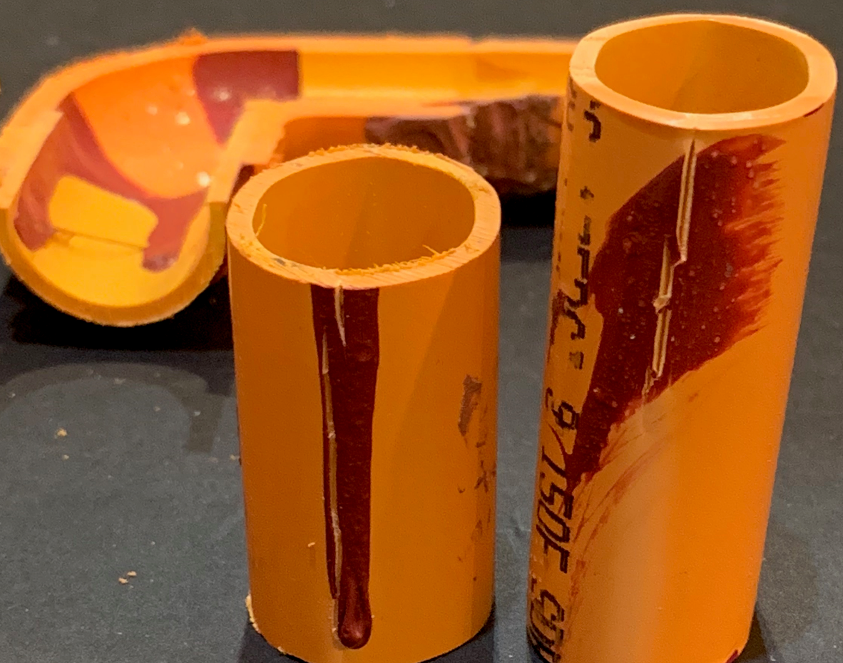 Sectioned orange CPVC pipe specimens and an elbow fitting on a dark bench, with dark solvent cement smeared and dribbled down the inside walls