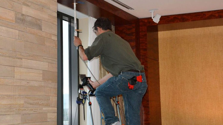 A worker on a ladder replacing white piping beside a window in a finished interior, with a ceiling sprinkler head visible above