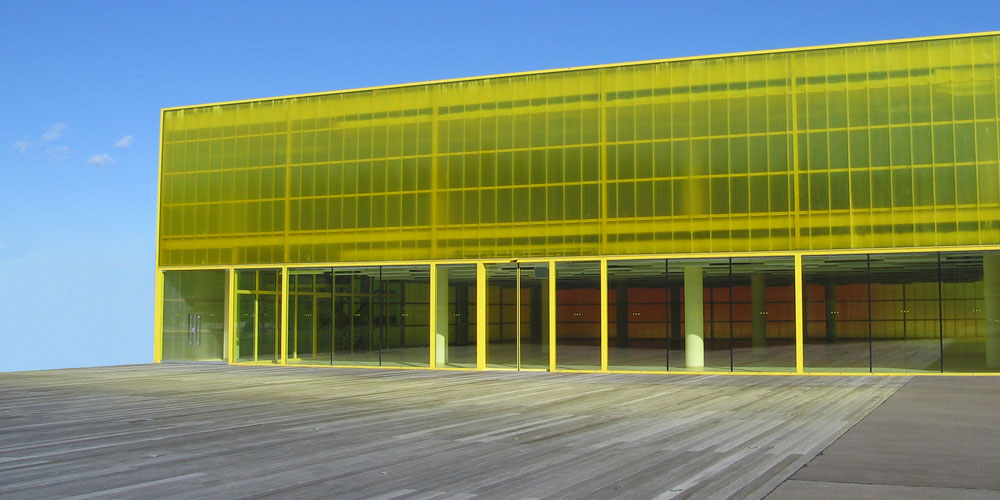 A modern building clad above its glazed ground floor in vivid, evenly coloured yellow translucent multiwall polycarbonate panels, photographed from a timber deck against a blue sky